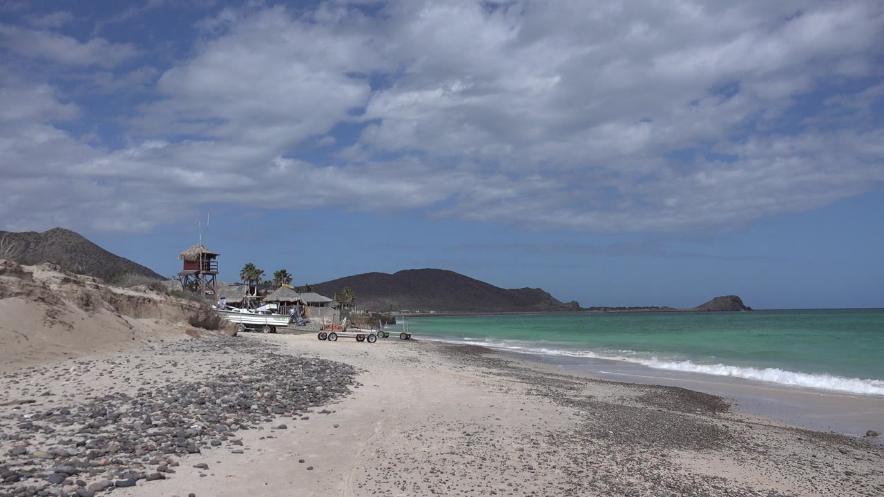 Cabo Pulmo shoreline one second in, showing the empty pebbled sand in the foreground, a thin line of surf, turquoise water, distant hills, and broad clouds overhead with no visible people.