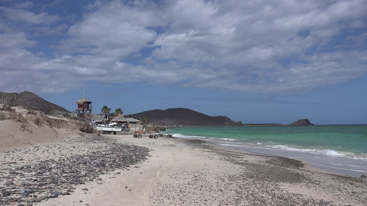 Opening view of a Cabo Pulmo beach with rocky sand in the foreground, turquoise-green water to the right, boats and a small shore structure at left, distant hills, and no visible people.