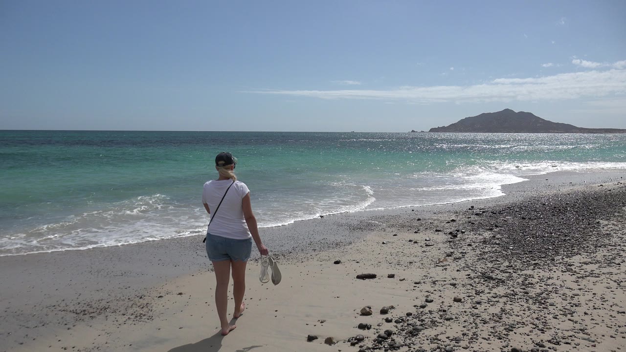 At 0:00 in Cabo Pulmo, Mexico, a barefoot woman in a white shirt and denim shorts walks toward turquoise surf while carrying shoes, with a low island or mountain on the horizon.