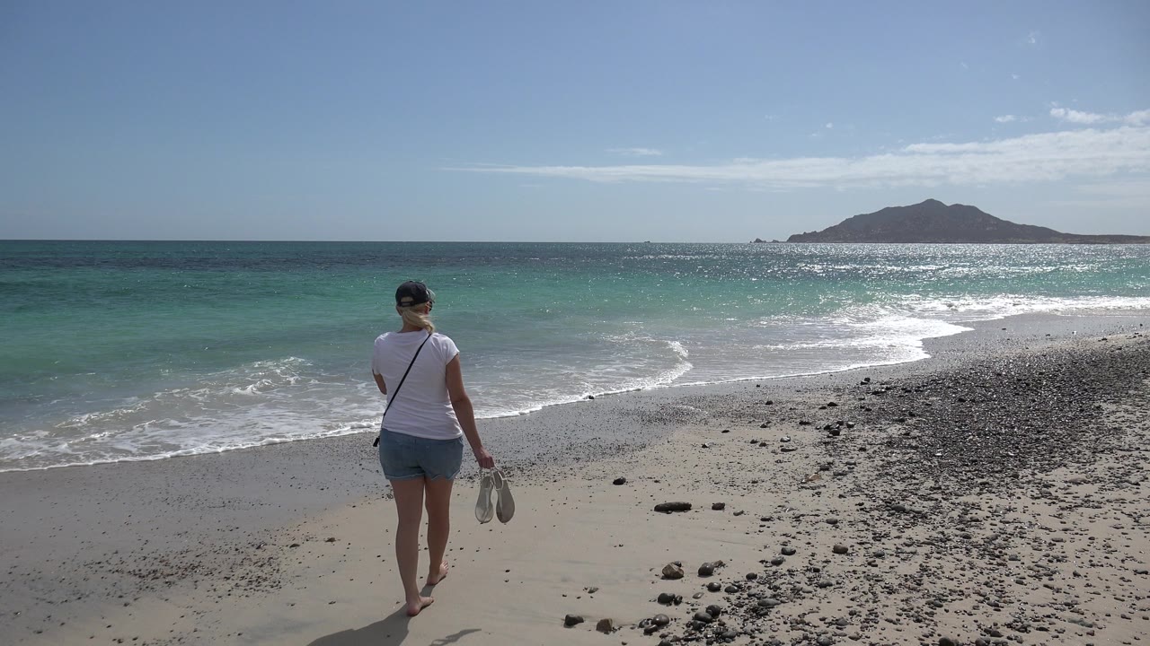 At 0:01 in Cabo Pulmo, Mexico, the woman stands closer to the waterline with her back to camera, shoes in one hand, as small waves curl across the sand.