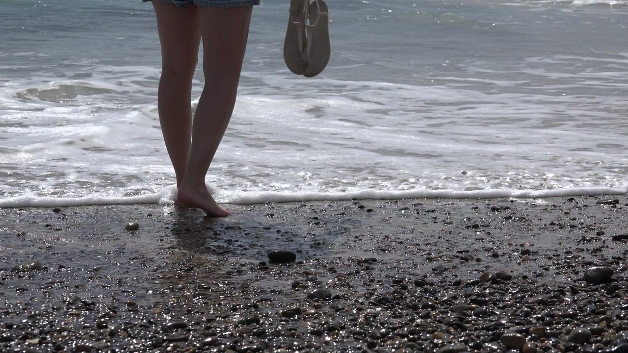 Bare legs at the edge of a pebbly beach with small waves behind.