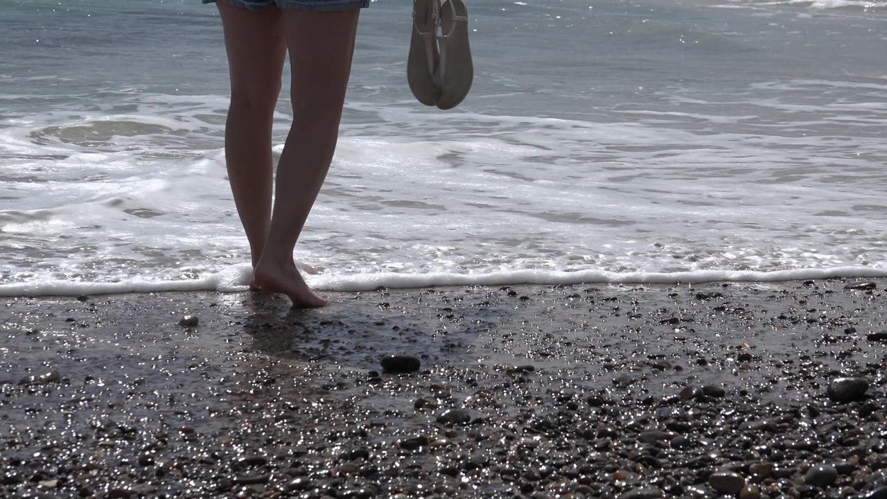 At 0:01 in Cabo Pulmo, Mexico, the same lower-body view shows the person facing the water while a narrow band of white foam moves across the wet pebbly sand.