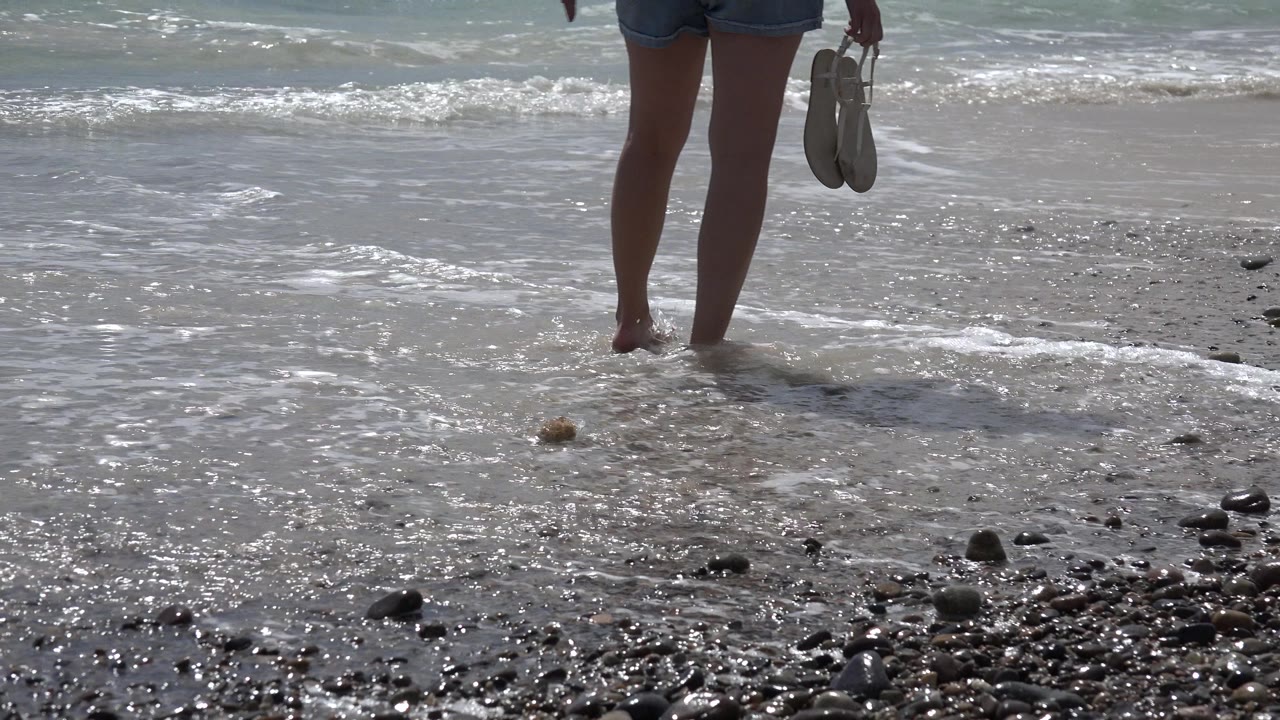 At Cabo Pulmo, Mexico, a cropped adult figure stands barefoot where a small wave reaches wet sand and scattered dark stones.