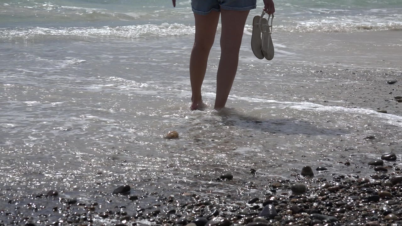 At Cabo Pulmo, Mexico, the cropped beach walker holds sandals while shallow foam spreads across wet sand dotted with rounded rocks.