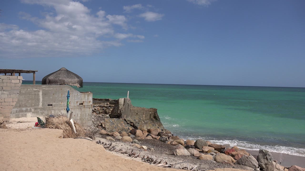 Sunny beach with concrete wall, thatched roof, rocks, and turquoise ocean.