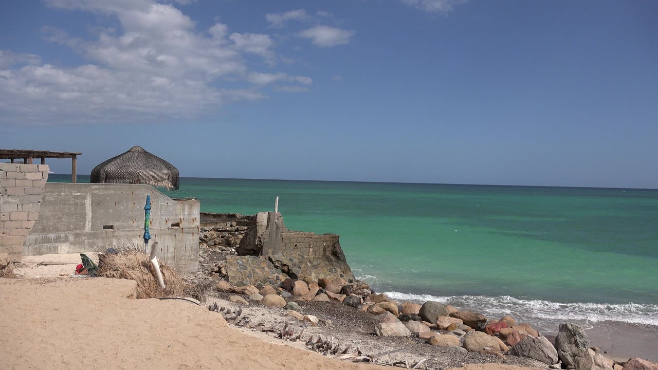 Opening frame in Cabo Pulmo, Mexico, showing pale sand, shoreline rocks, turquoise water, a low concrete structure, a thatched roof, and no visible people.