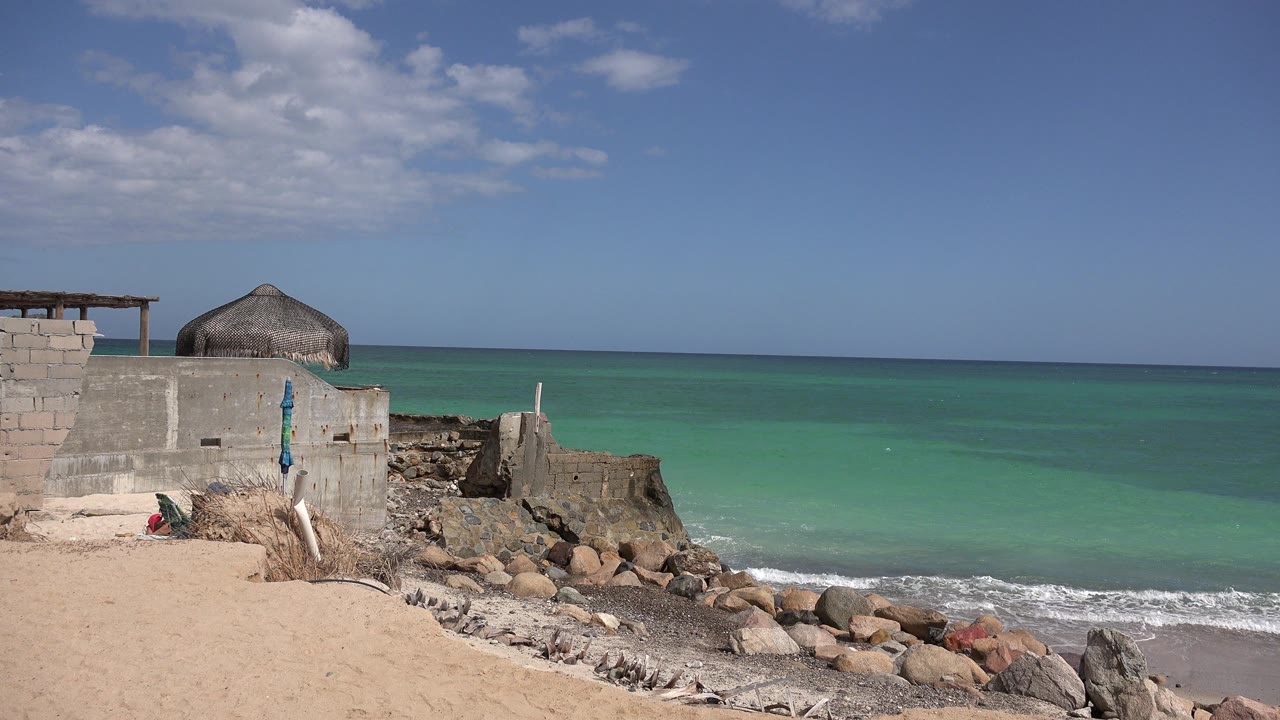 At 0:01 in Cabo Pulmo, Mexico, small wave foam reaches the rocks below the concrete shore structure, with open turquoise water and no visible people.