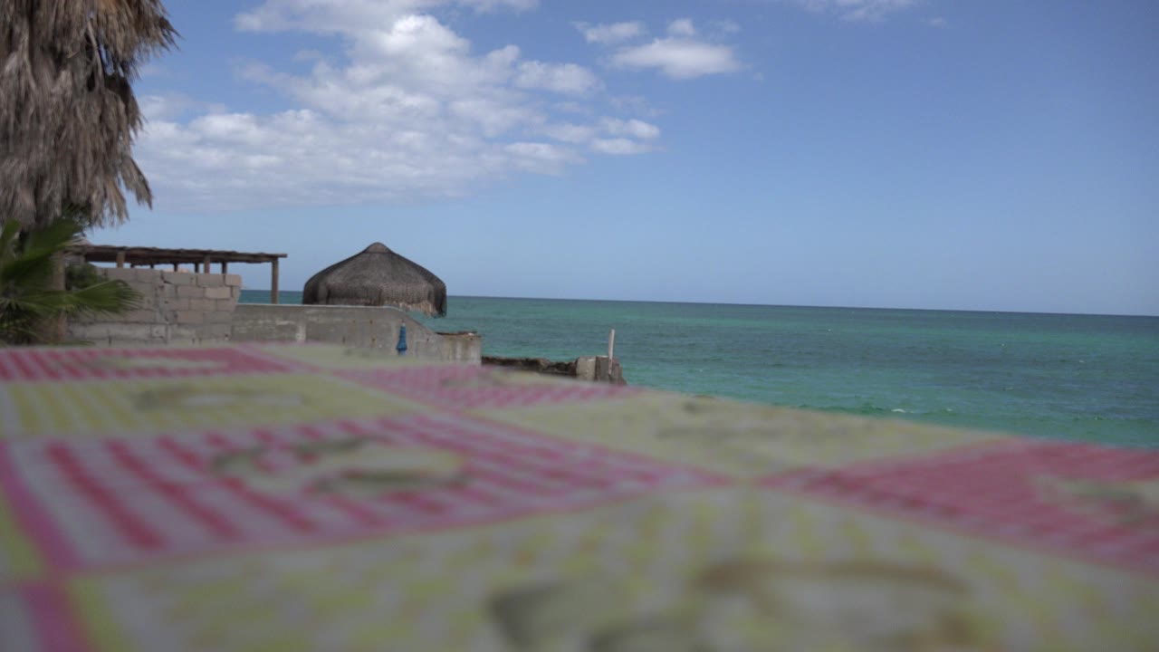 Cabo Pulmo coastal view at the start of the clip, with blurred striped fabric in the foreground, a low wall, a thatched roof structure, blue water, and no visible people.