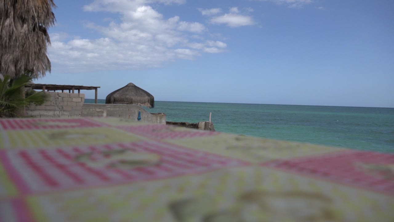 Blurred towel foreground with ocean, palm fronds, low wall, and thatched hut beyond.