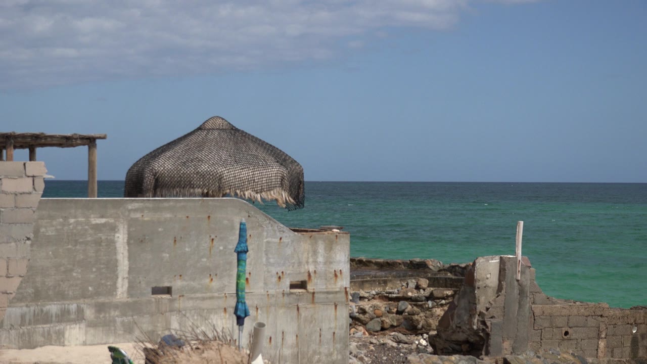 Opening frame of Cabo Pulmo construction by the sea, with a gray concrete wall, thatched roof, rubble, blue post, turquoise water, and no visible people.