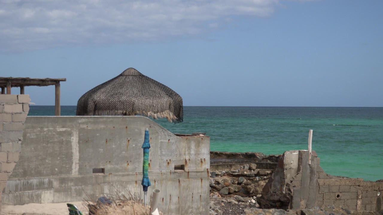 At 0:00, a thatched cabana roof sits behind a rough cinder block wall on the Cabo Pulmo coast, with turquoise water and no visible people.