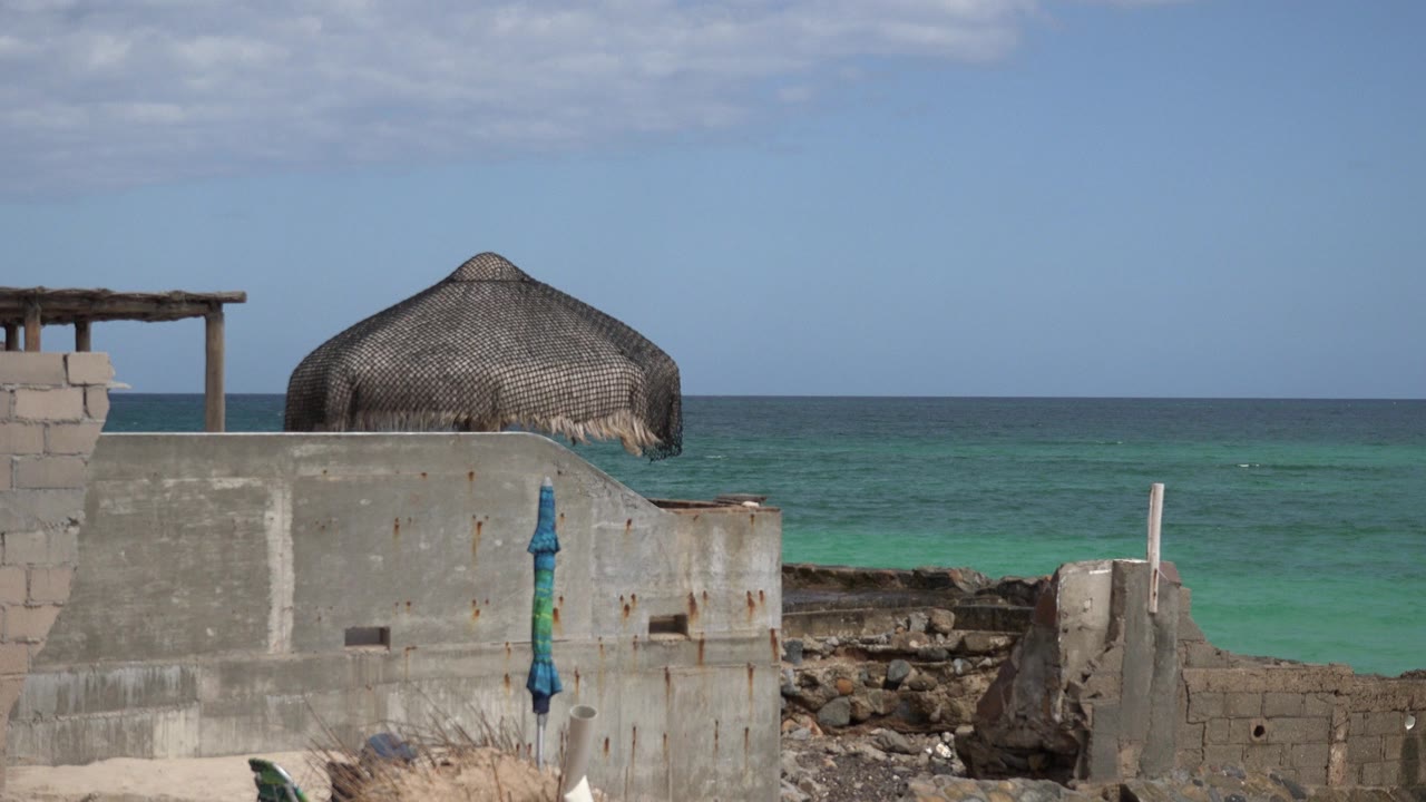 At 0:01, the cabana roof remains centered behind worn masonry in Cabo Pulmo, with a straight ocean horizon and no visible people.