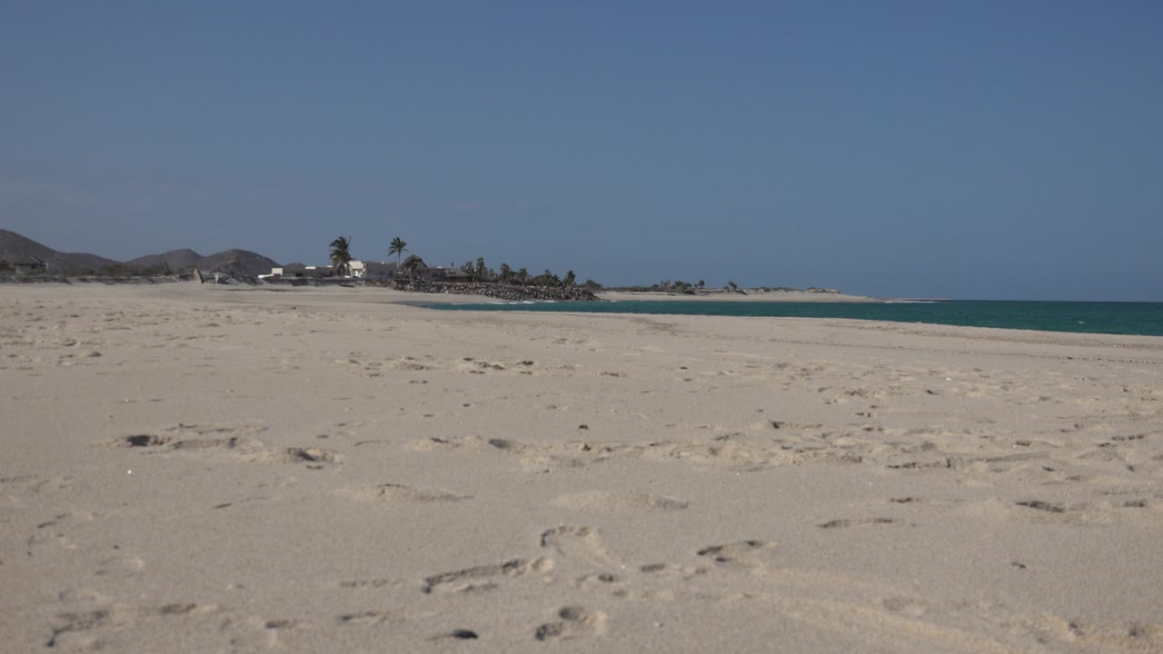 Cabo Pulmo beach view at the start of the clip, with footprint-marked sand in front, blue sea to the right, palm trees and low hills in the distance, and no visible people.