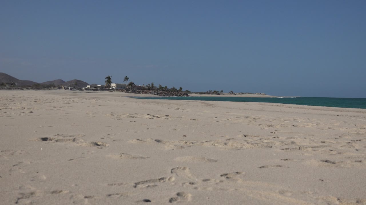 Opening frame of a Cabo Pulmo beach with pale sand and footprints in the foreground, distant palms and buildings near the shore, blue water at right, and no visible people.