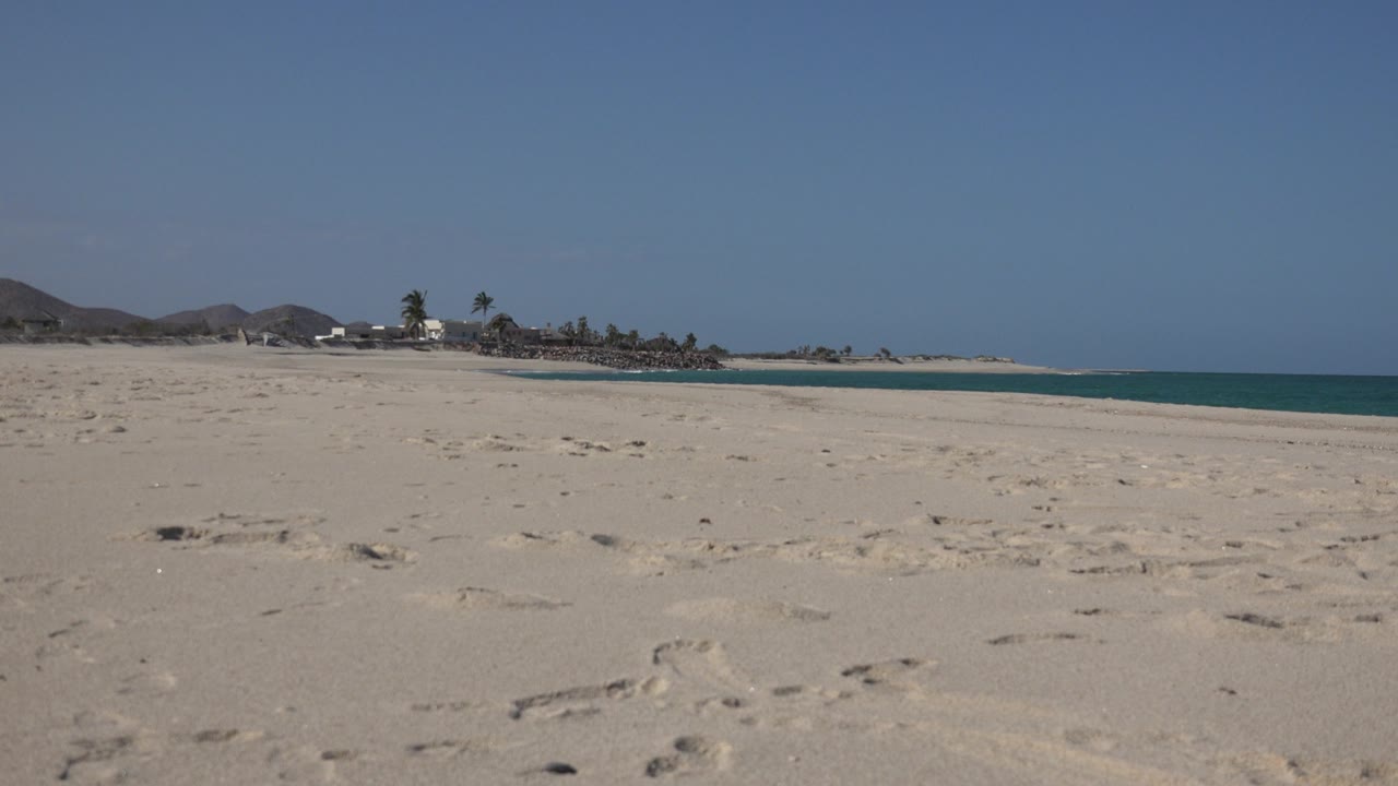A wide Cabo Pulmo shoreline view shows textured sand and footprints across the foreground, turquoise water along the right edge, distant palms, low hills, and no visible people.