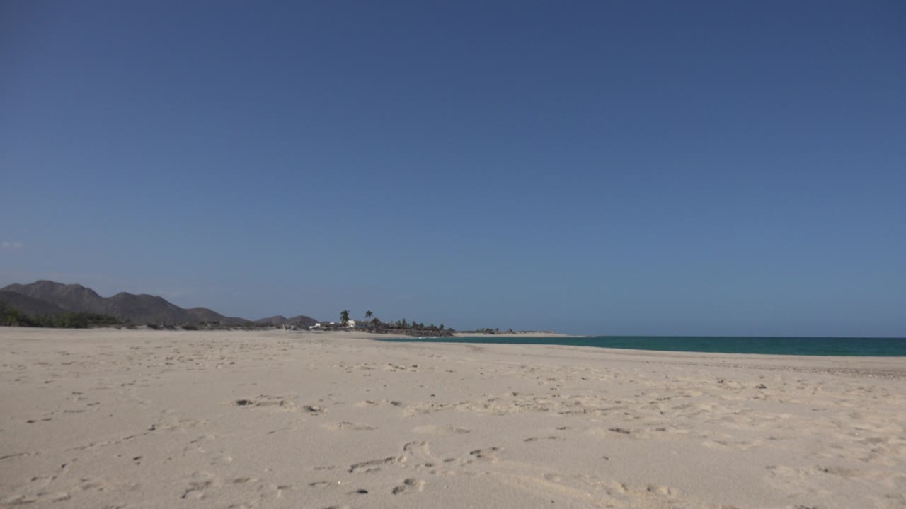 Opening view of a wide empty beach in Cabo Pulmo, Mexico, with pale sand in front, blue ocean to the right, distant low mountains, a small shoreline building, and no visible people.