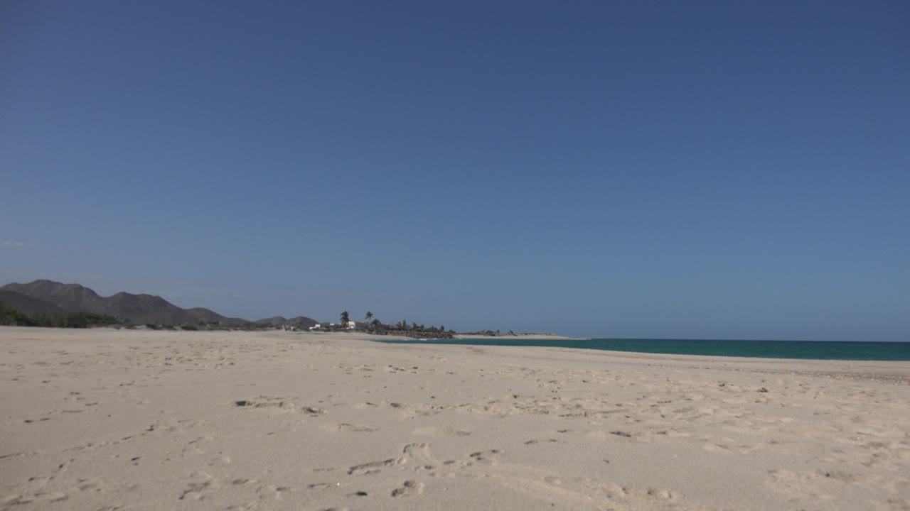 Cabo Pulmo beach seen from low on the sand with distant mountains at left, turquoise water at right, bright blue sky overhead, and no visible people.