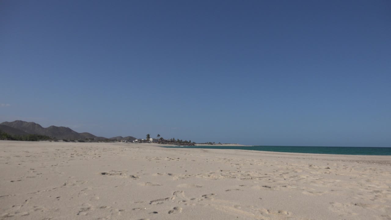 Cabo Pulmo beach remains open and uncrowded, with textured sand in front, low hills and small structures in the distance, ocean to the right, and no visible people.
