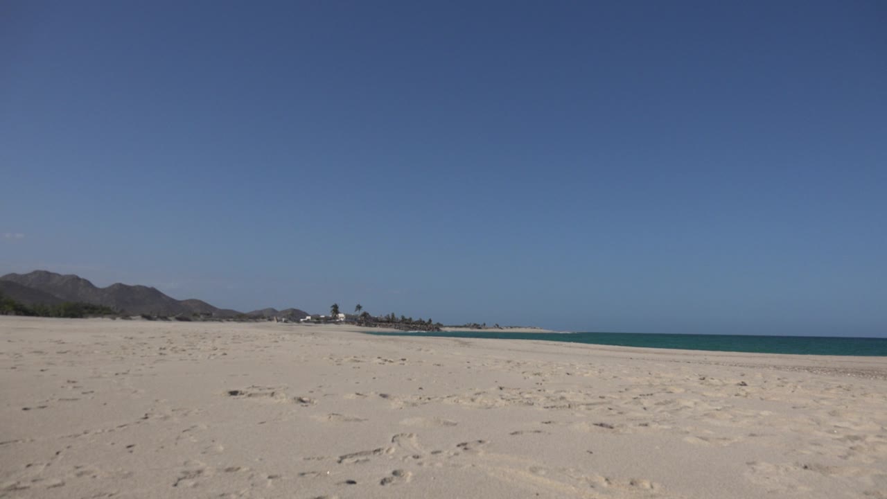 Opening frame of a wide Cabo Pulmo beach view with pale sand, small rocks, distant low mountains, a thin blue waterline, and no visible people.