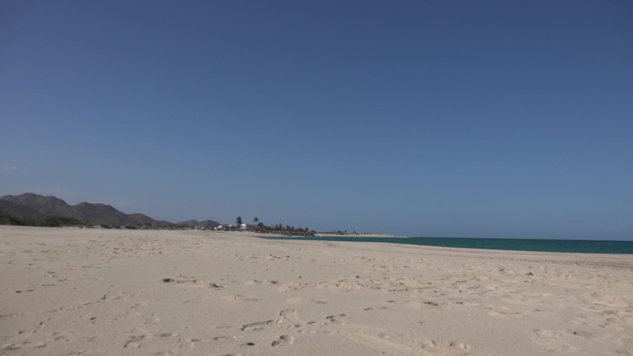 Cabo Pulmo beach frame with footprint-marked sand in front, distant palms and buildings near the left horizon, ocean on the right, and no visible people.