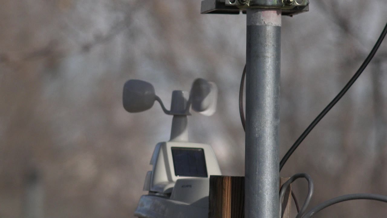 A Westminster, Colorado weather station with cup-style wind sensor and dark solar panel is partly framed by a metal pole, with blurred bare trees behind it and no visible people.