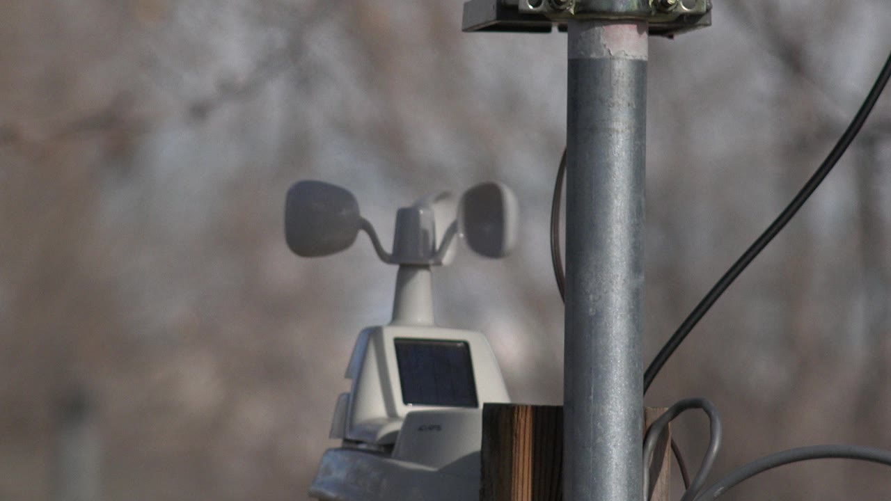 The Westminster weather station remains centered near the metal pole, with the left wind cup in shadow, the solar panel below, soft gray-brown trees in the background, and no visible people.