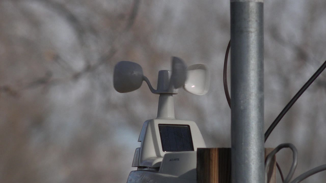 A small weather sensor with wind cups and a dark solar panel is framed beside a vertical metal pole in Westminster, Colorado, with no visible people.