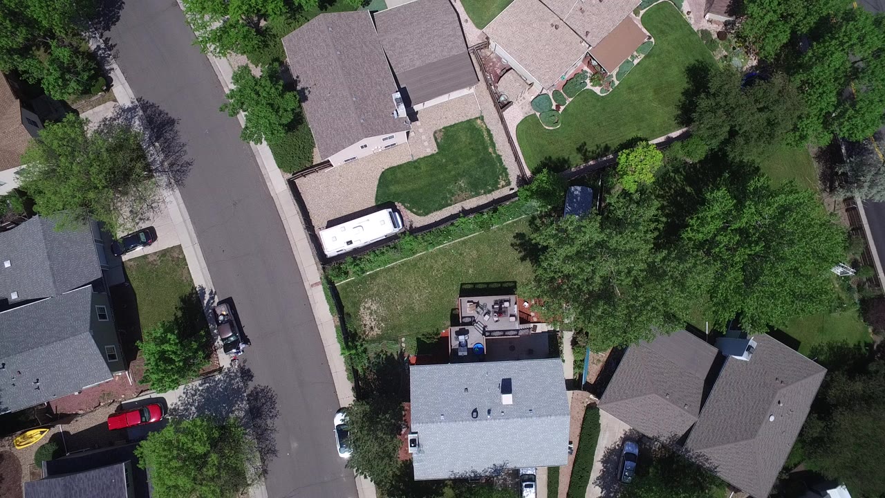 Overhead view in Westminster, Colorado showing a curved residential street, roofs, green yards, mature trees, a white RV, and parked cars with no visible people.