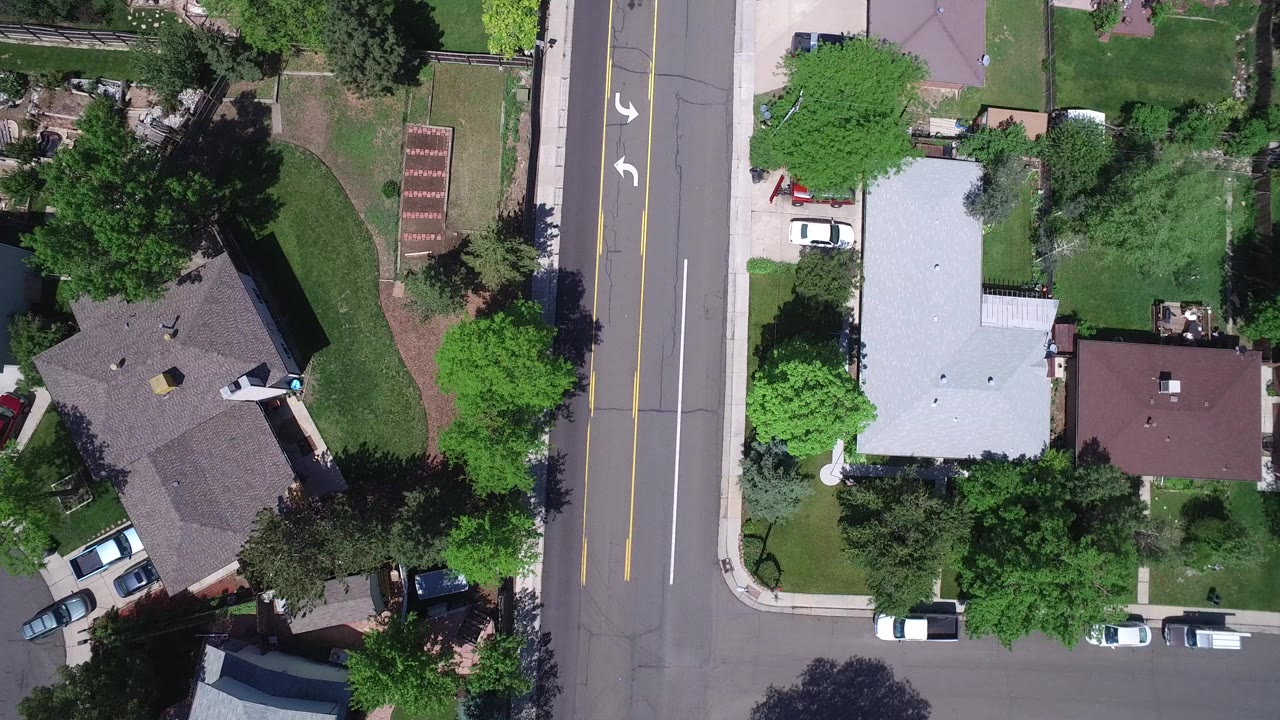 Overhead view of houses, lawns, trees, parked cars, and a residential road.