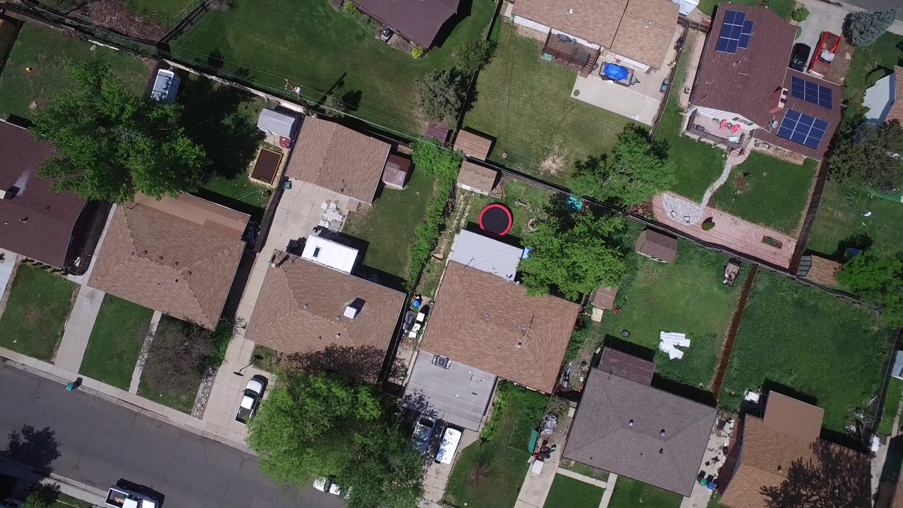 Aerial frame over Westminster, Colorado showing tan and brown rooftops, fenced yards, tree cover, a residential road, driveways, parked vehicles, solar panels, and no visible people.
