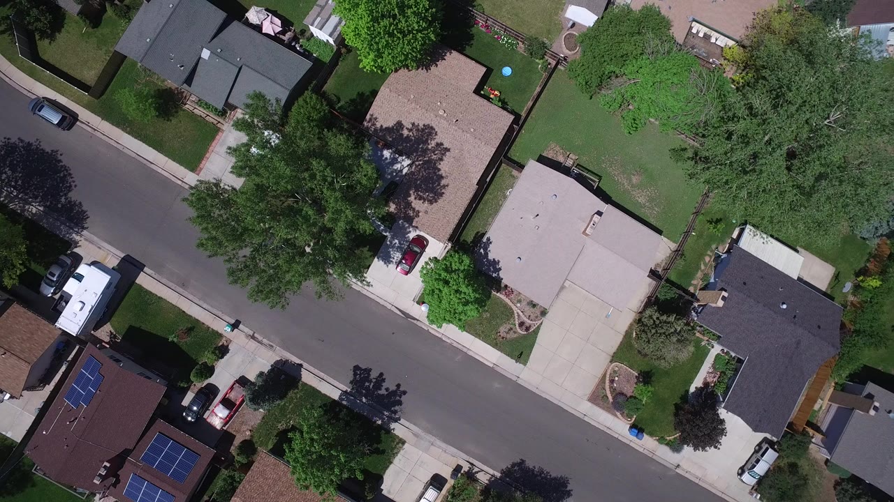 Overhead suburban street with homes, lawns, trees, and parked vehicles.