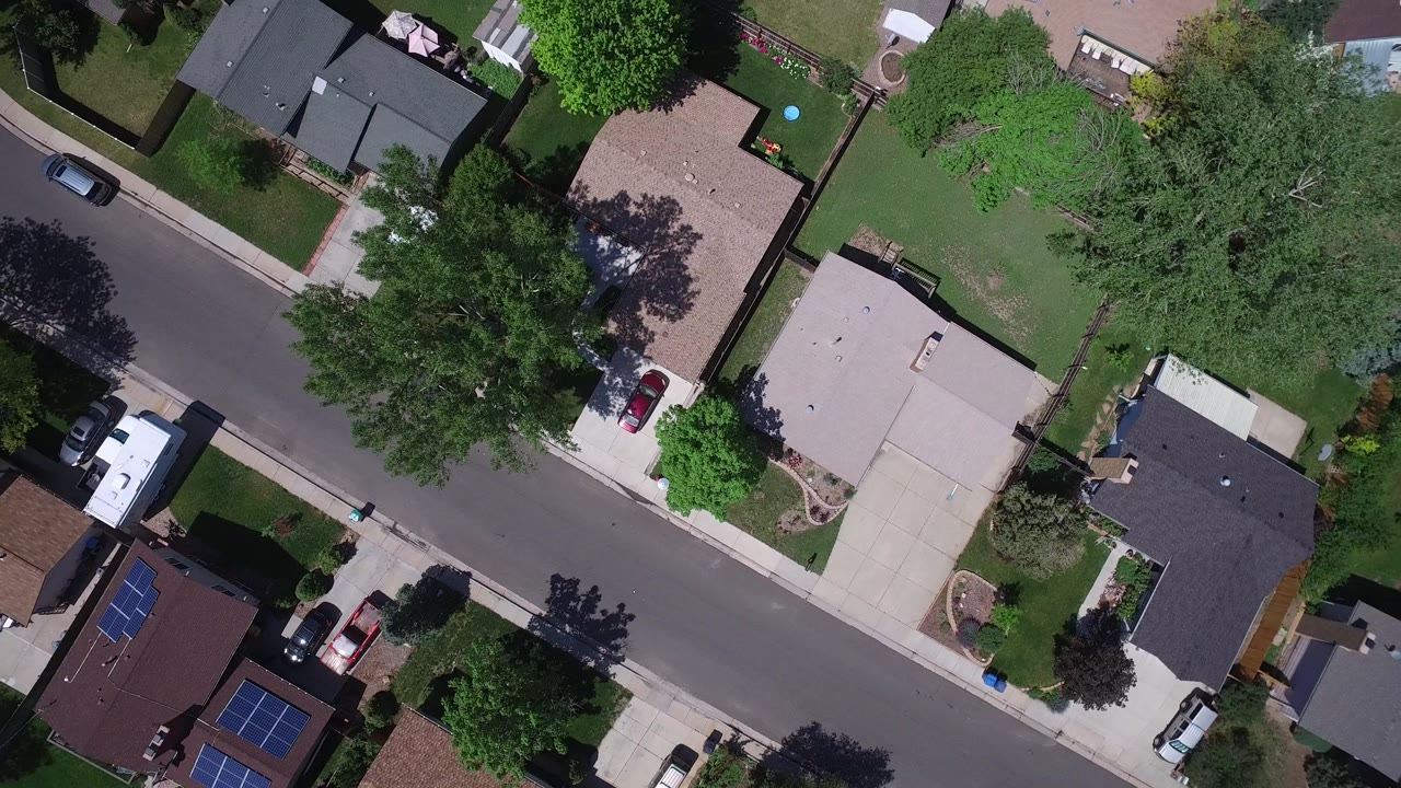 Overhead Westminster, Colorado frame with an asphalt street crossing between rooftops, driveways, shaded yards, trees, and several parked cars, with no clearly visible people.
