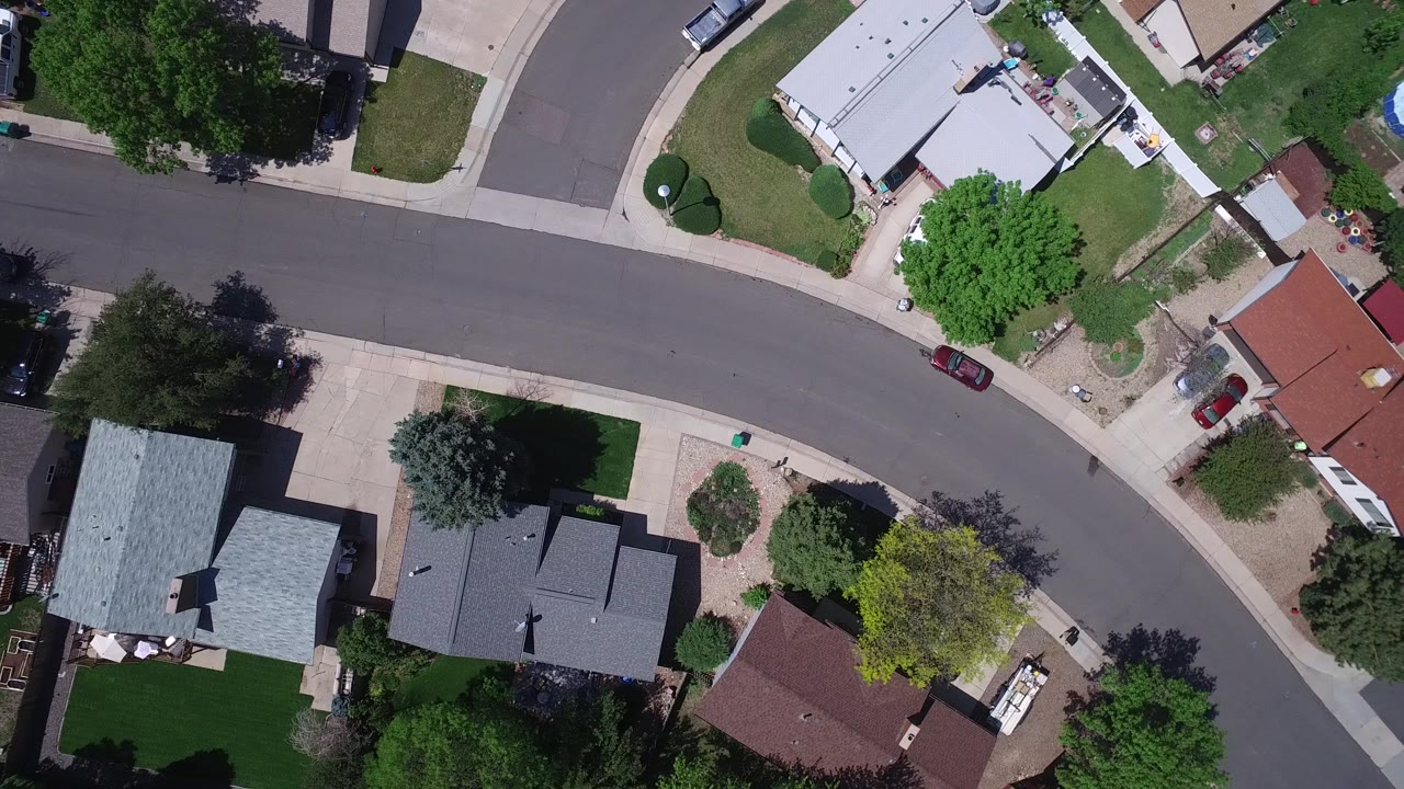 Aerial frame in Westminster, Colorado showing the curved road centered between detached homes, green yards, large trees, sidewalks, and no clearly visible people.