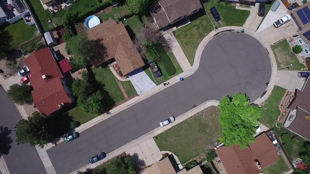 Aerial frame over Westminster, Colorado homes with the cul-de-sac centered, a white car along the curb, green lawns, trees, and no visible people.