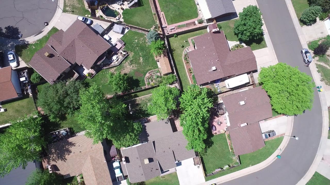 Overhead aerial frame in Westminster, Colorado showing detached homes, green lawns, driveways, parked cars, and a curving residential street, with no visible people.