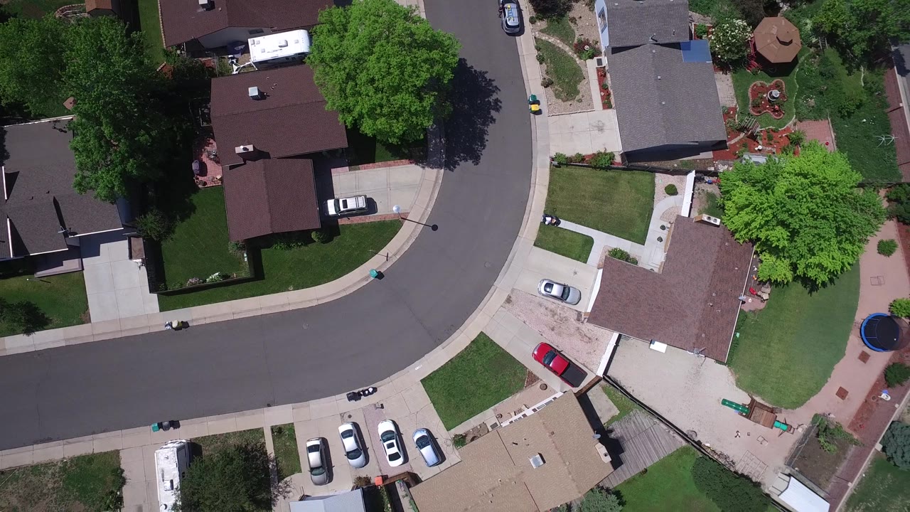 Aerial subdivision view with homes, a red car, lawns, and a larger road at the edge.