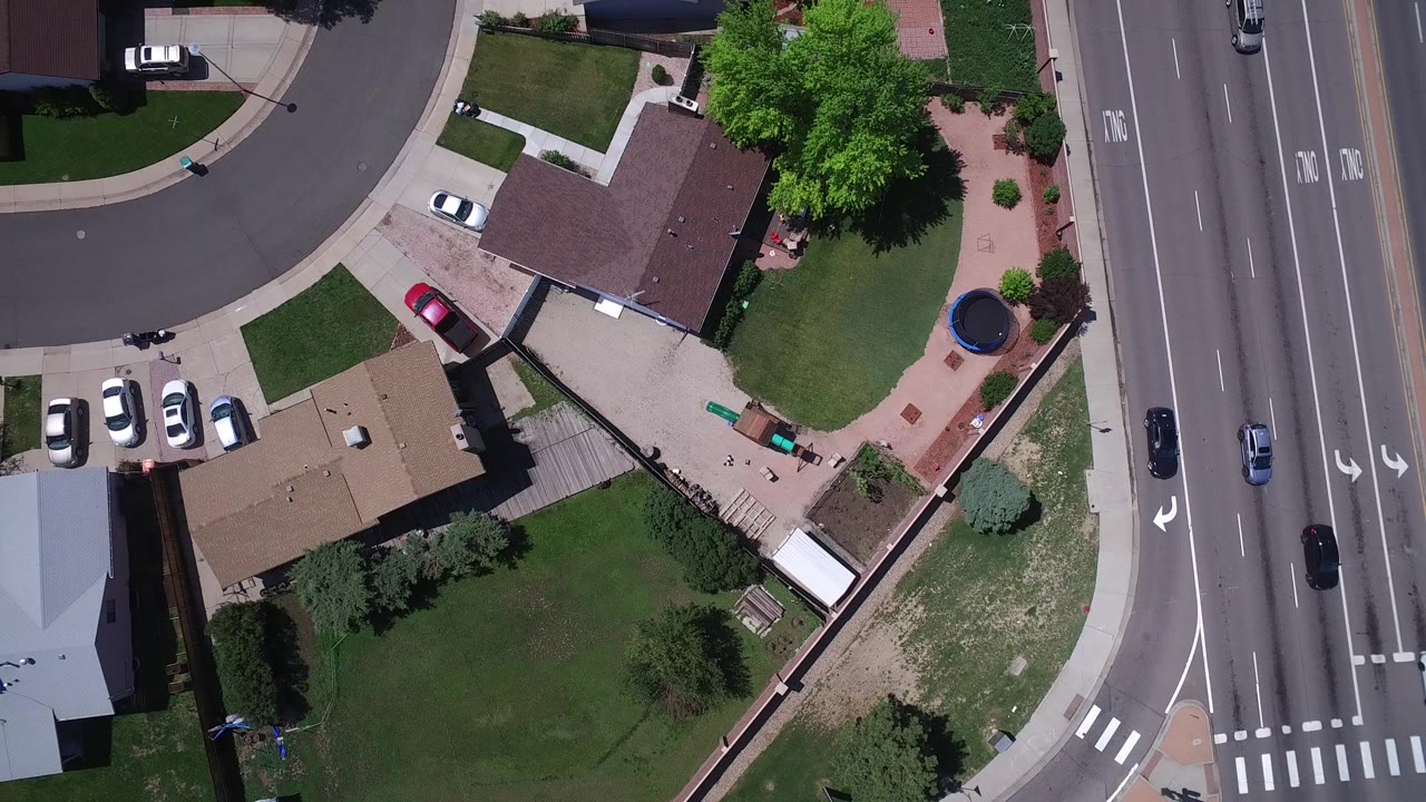 Top-down aerial frame over Westminster, Colorado showing residential rooftops, fenced yards, green lawns, a curved side street, and a multi-lane road with cars, with no visible people.