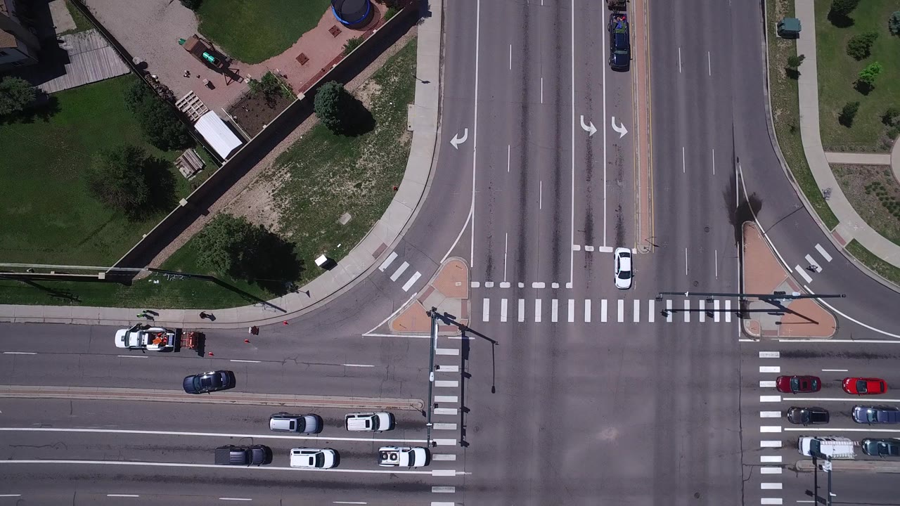Aerial frame over Westminster, Colorado showing cars in several lanes, a red car near the lower roadway, broad crosswalk markings, traffic light poles, sidewalks, and small visible pedestrians.