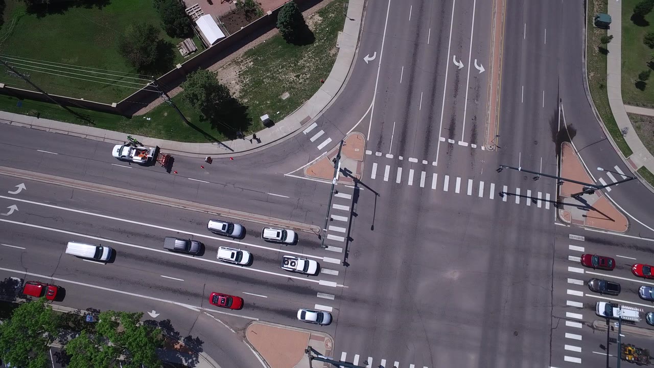 Top-down road intersection with cars, crosswalks, lawns, and trees.