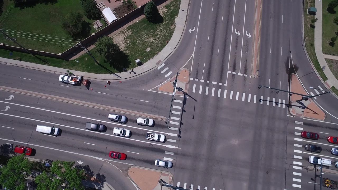 Top-down Westminster, Colorado road scene with a white marked vehicle near the upper-left curb, cars moving through the lower lanes, crosswalks, sidewalks, grass, and no clearly visible people.