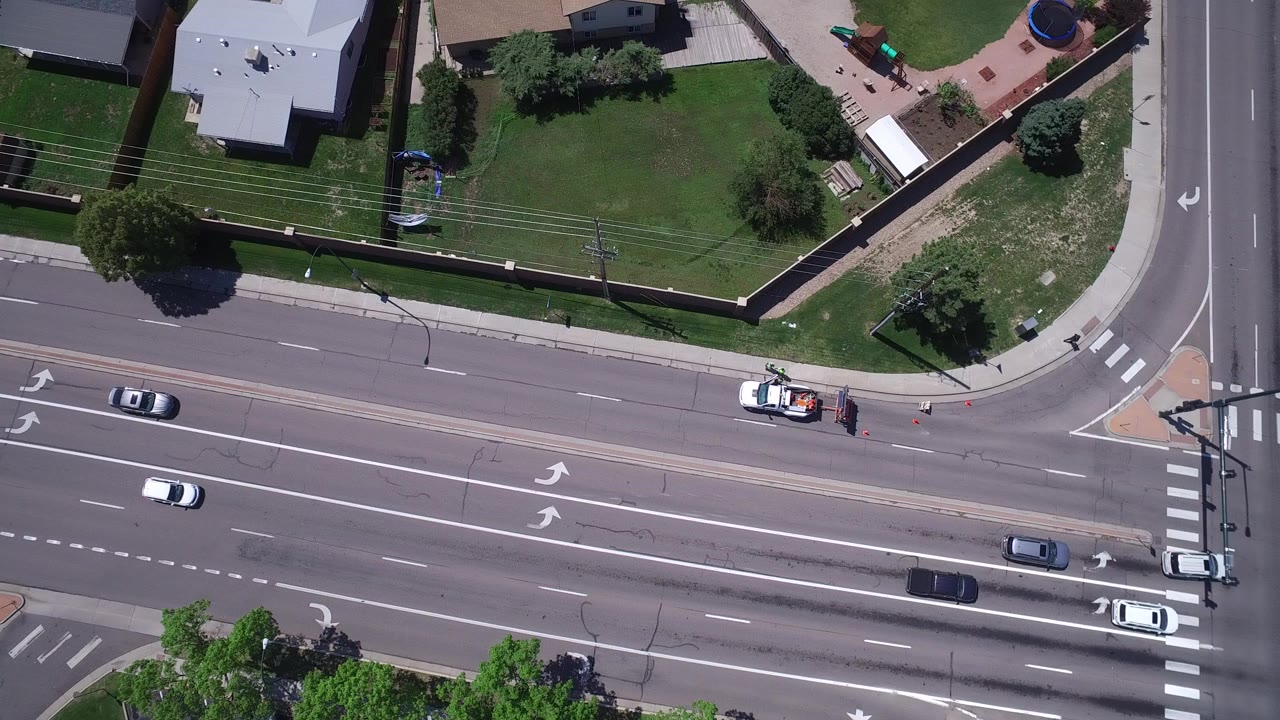 Overhead frame from Westminster, Colorado showing several cars on a multi-lane road, fenced yards, lawns, trees, sidewalks, and no visible people.