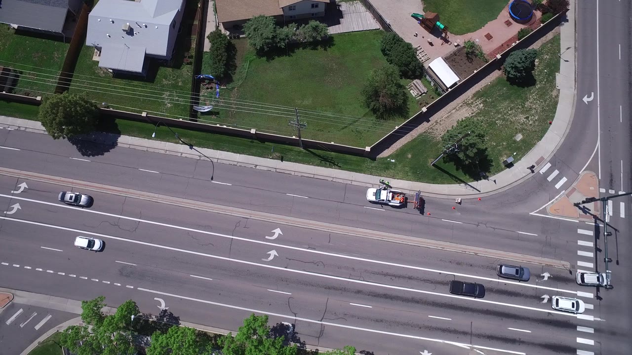 Westminster, Colorado aerial frame with cars spaced across the lanes, a small roadside work vehicle near an intersection, fenced homes, green lawns, and no visible people.