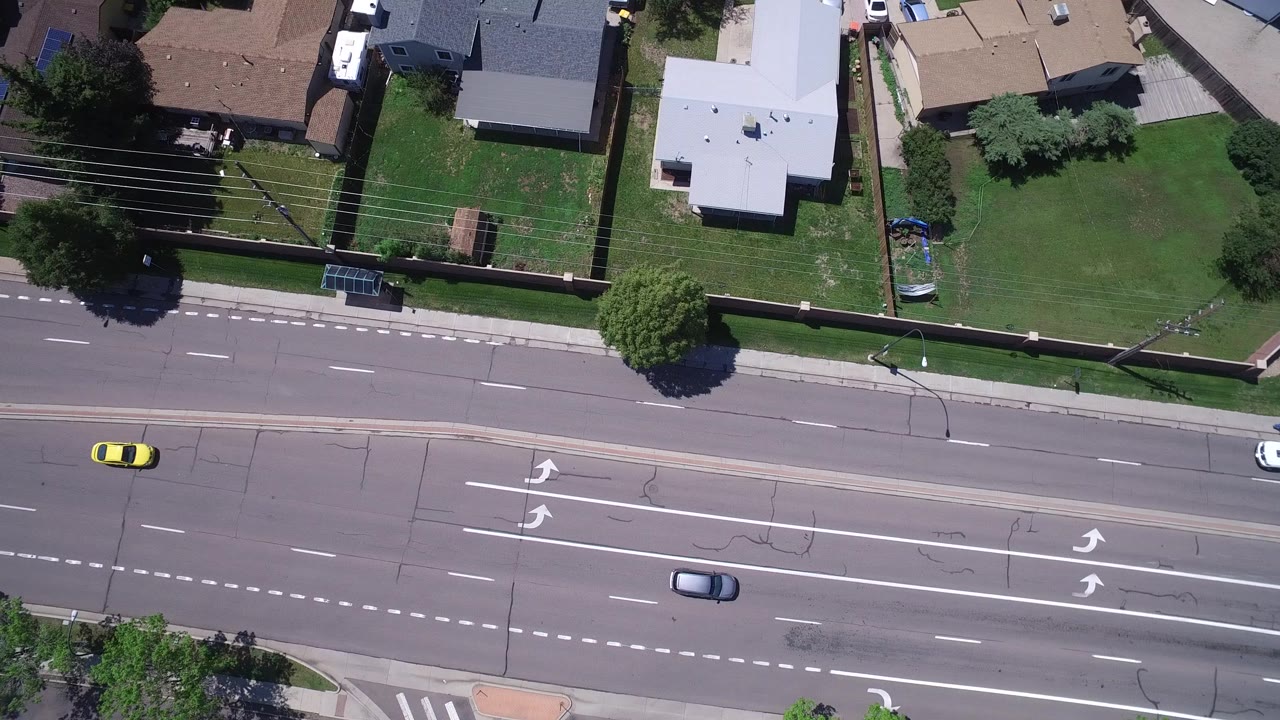 Overhead Westminster, Colorado frame with a yellow car and a white car on the road, residential yards and rooftops above the street, and no visible people.