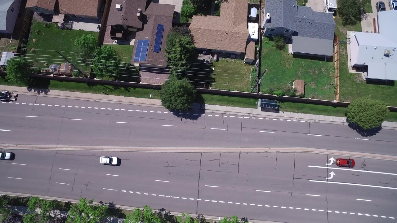 Overhead frame in Westminster, Colorado showing a wide multi-lane road, moving cars, sidewalks, lawns, and private houses, with no visible people.
