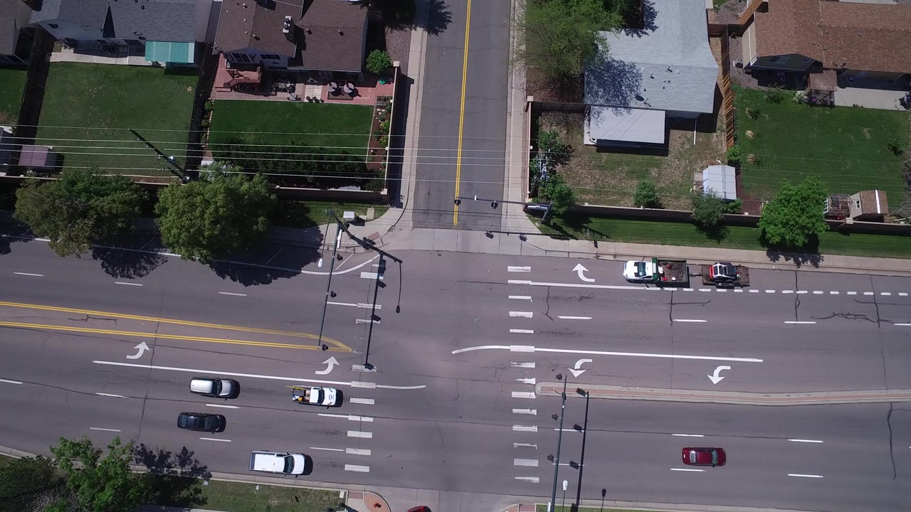 Top-down aerial frame in Westminster, Colorado showing a broad avenue, a perpendicular residential street, crosswalks, lawns, rooftops, several cars, and no visible people.