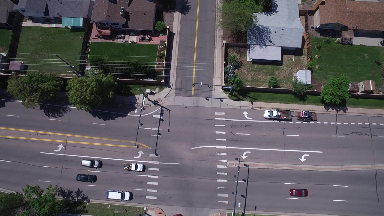 Aerial Westminster, Colorado frame with cars spaced across the avenue, white turn arrows, crosswalk stripes, a center median, residential yards, and no visible people.