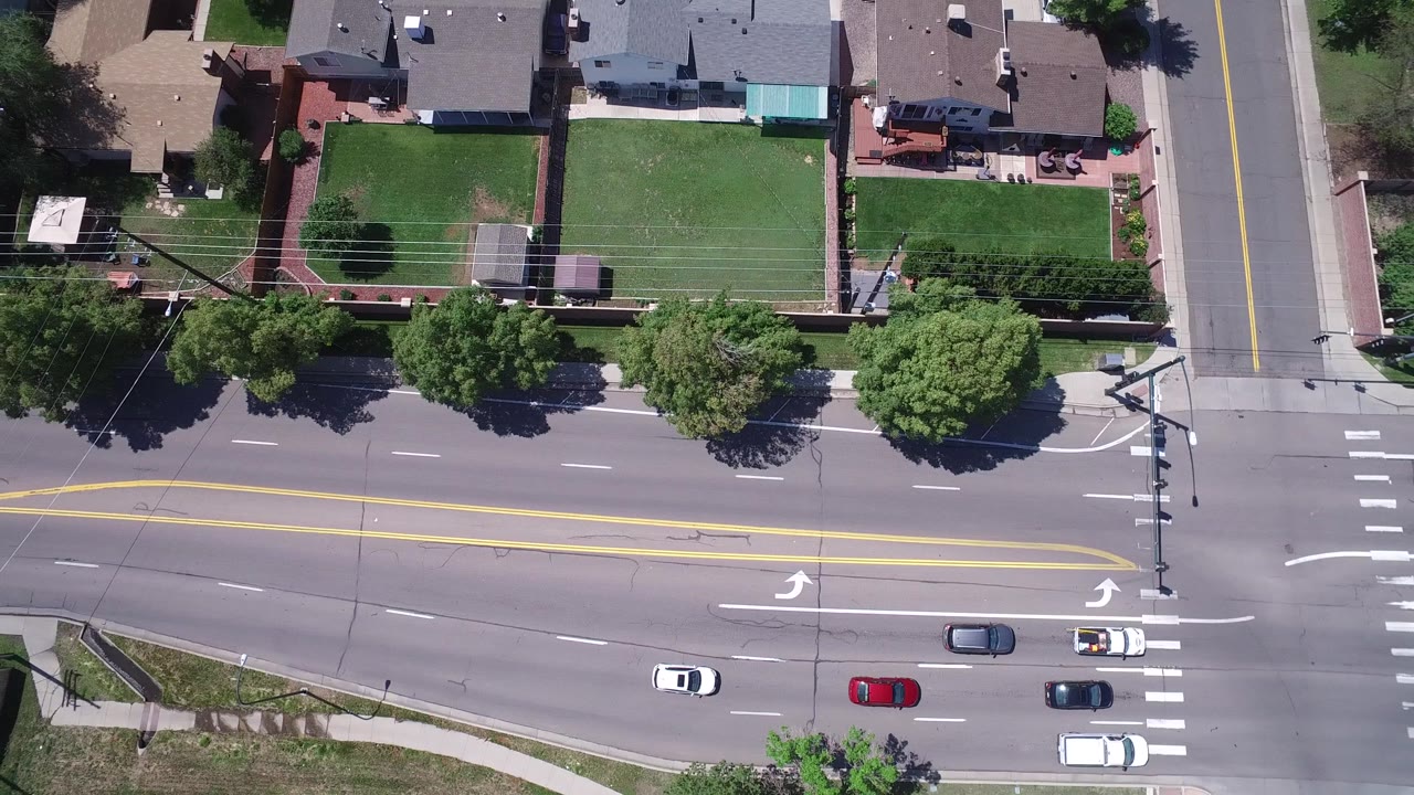 Aerial frame over Westminster, Colorado with several cars on the paved road, yellow center lines, turn arrows, fenced green lawns, mature trees, and detached homes with no visible people.