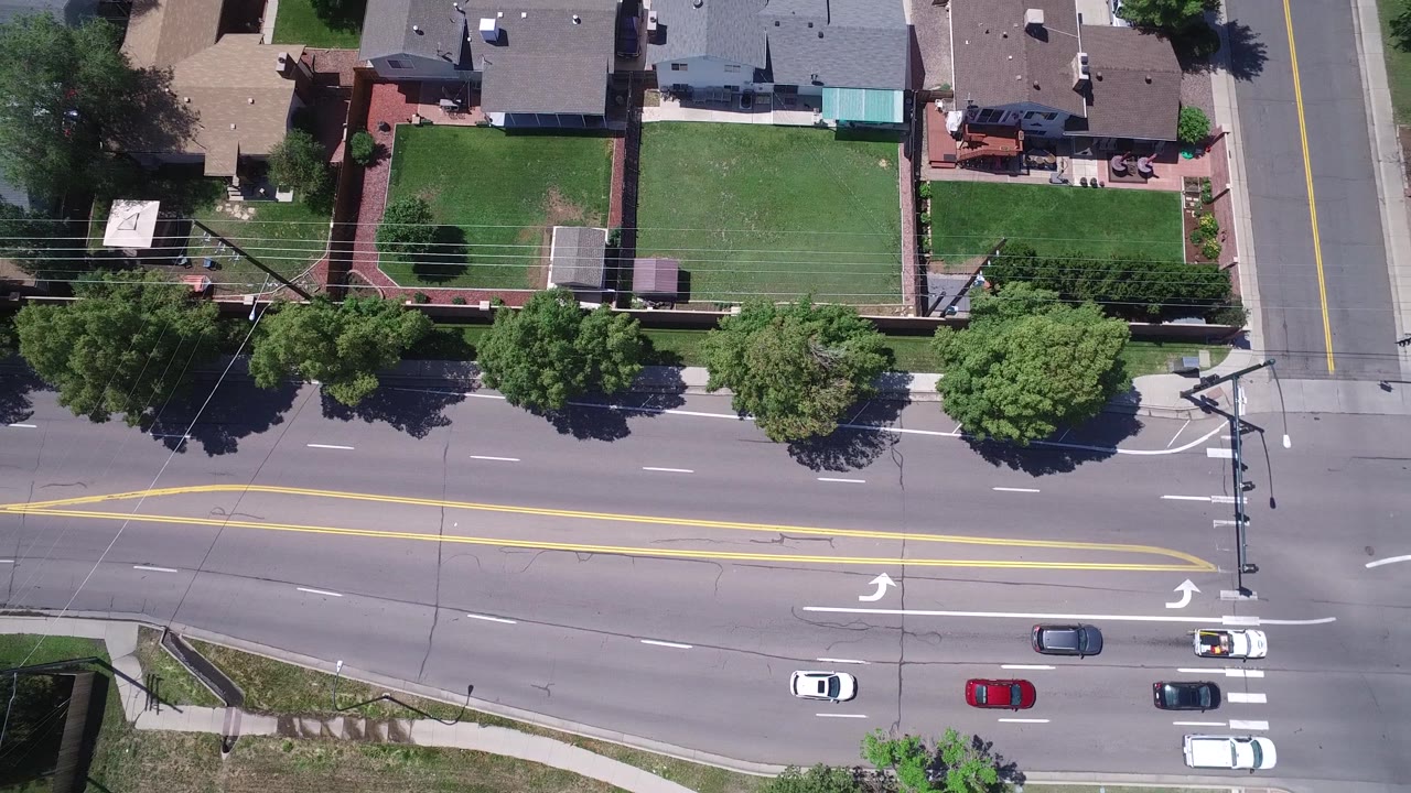 Top-down Westminster, Colorado frame showing cars spaced across the roadway, a side-street intersection, white crosswalk bars, green residential yards, fences, trees, and rooftops with no visible people.