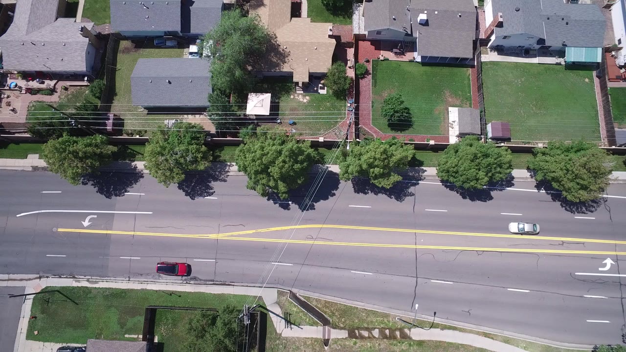 Straight-down aerial frame in Westminster, Colorado showing a multi-lane road, yellow turn-lane markings, detached homes, lawns, trees, fences, and no clearly identifiable people.