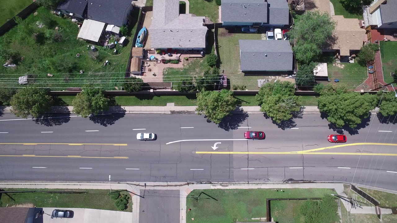 High aerial view in Westminster, Colorado showing a multi-lane road, sidewalks, lawns, trees, rooftops, and several cars, with no visible people.
